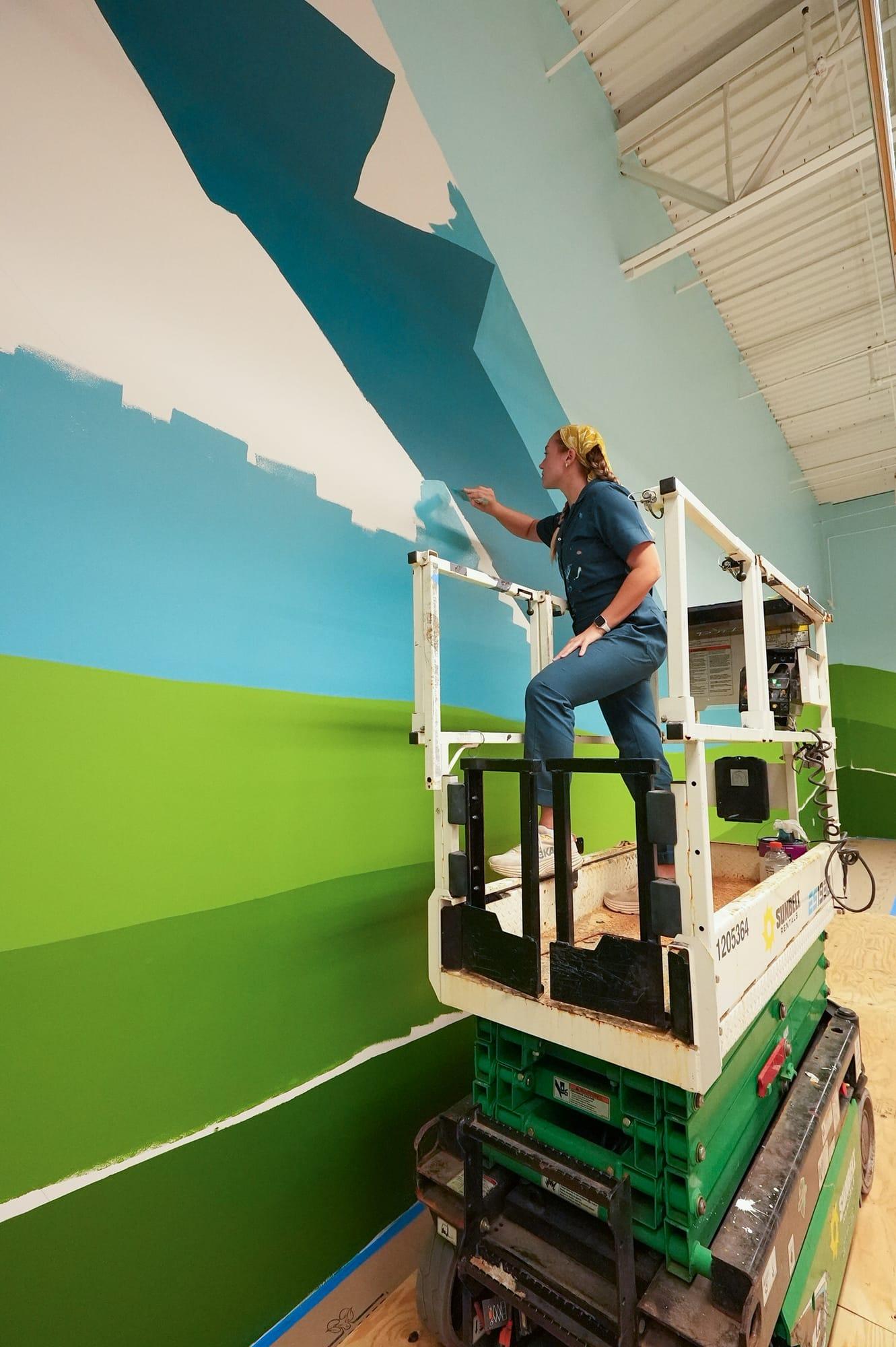 Tori on a scissor lift hand-painting a large landscape mural across a commercial wall
