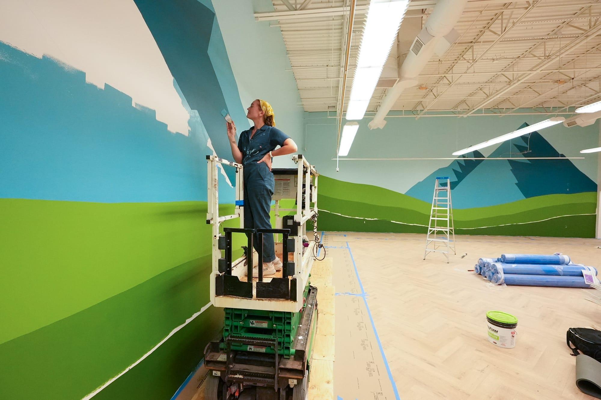 Tori painting a mountain landscape mural from a scissor lift inside a large commercial space