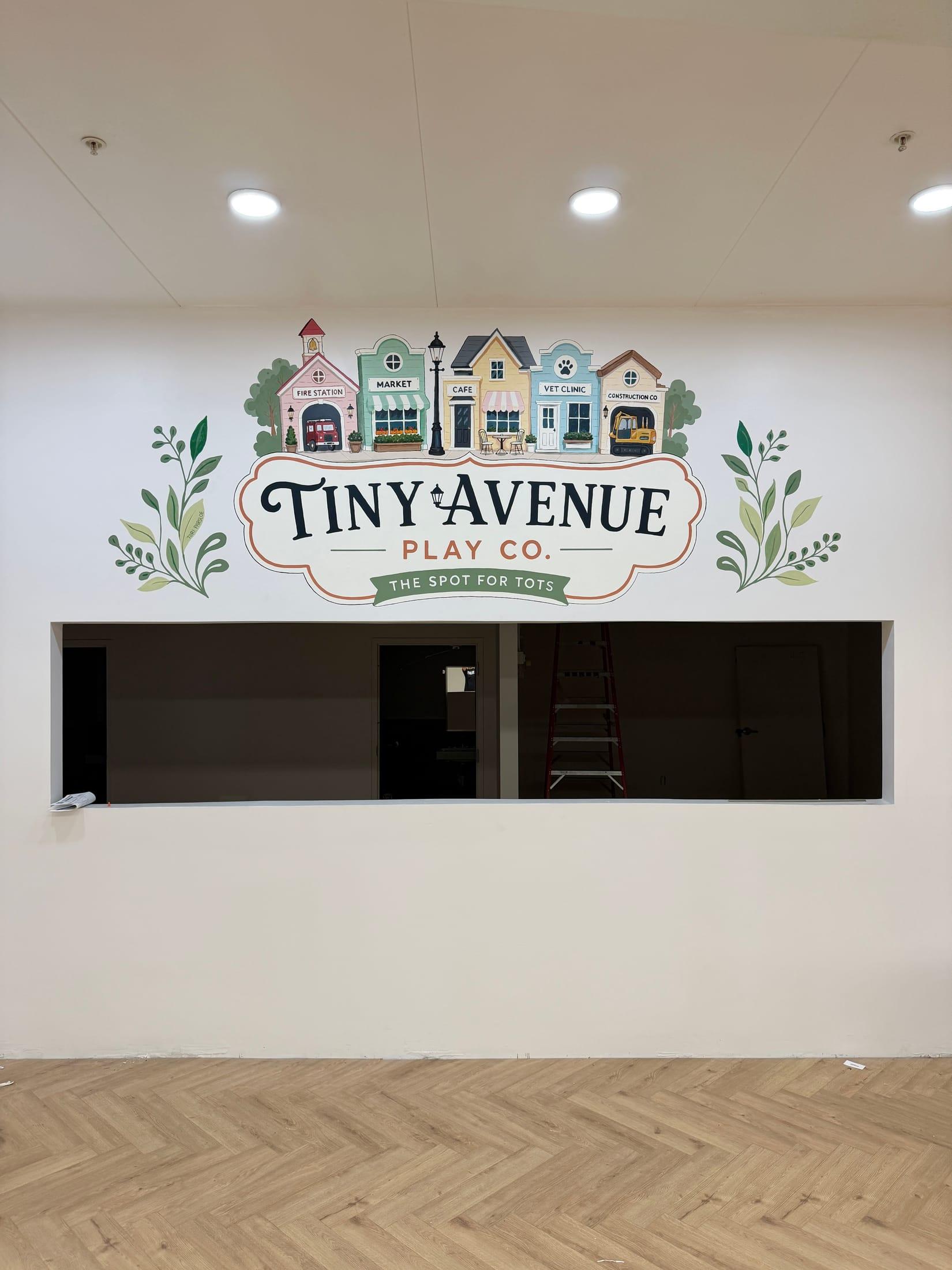 Hand-painted Tiny Avenue Play Co. mural with illustrated storefronts above a window in a children's play space